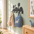 Zwarte dinosaurus-kapstok met een blauw kinderjasje en crème rugzak naast een houten bed in een lichte dino-kinderkamer.