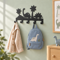 Zwarte metalen dino kinderkapstok met vijf haken aan een saliegroene muur, met een crème vest en blauwe dinosaurus-rugzak.
