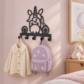 Zwarte metalen eenhoornkapstok aan een zachtroze muur, met een crème vest en lila kinderrugzak.